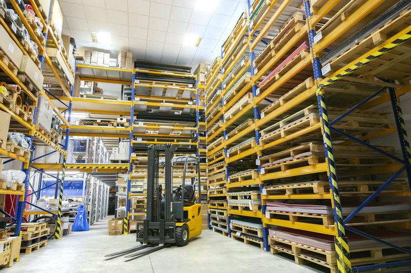 Interior de un almacén industrial con racks metálicos azules y amarillos, tarimas de madera apiladas y un montacargas amarillo en el pasillo